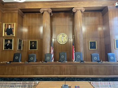 Seven seats sit at a raised desk. They sit in front of a large Classical-Revival style wall. Portraits hang on the walls of the room.