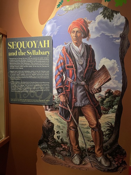 Sequoyah. He is depicted wearing a brightly-colored jacket and holding a book and walking cane. He is wearing a hair wrap, and looks to be about 40.