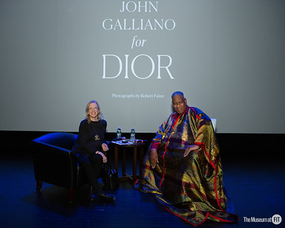 Talley and Steele are both seated. Talley, right, is wearing a patterned caftan, and Steele is wearing all black. Behind them is a large screen that reads, “John Galliano for Dior.”