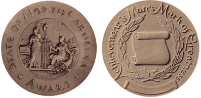 Image of the front and back of the North Carolina Award medal. The medal is gold in color and composition and has raised images and text. The front reads "State of North Carolina Award" with the state seal, an image of two women in robes representing Liberty and Plenty, one holding a capped pole and one holding heads of grain with a cornucopia. The back of the medal says "Achievement is Man's Mark of Greatness" and has an unfurling scroll surrounded by 