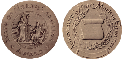 Image of the front and back of the North Carolina Award medal. The medal is gold in color and composition and has raised images and text. The front reads &quot;State of North Carolina Award&quot; with the state seal, an image of two women in robes representing Liberty and Plenty, one holding a capped pole and one holding heads of grain with a cornucopia. The back of the medal says &quot;Achievement is Man's Mark of Greatness&quot; and has an unfurling scroll surrounded by leafy branches.
