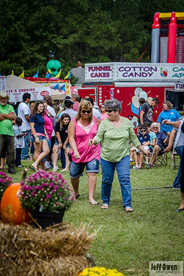 People gathered outside at a festival. There are tents with vendors and food trucks around. Flowers and pumpkins sit on haybales in the foreground and there are trees in the background.