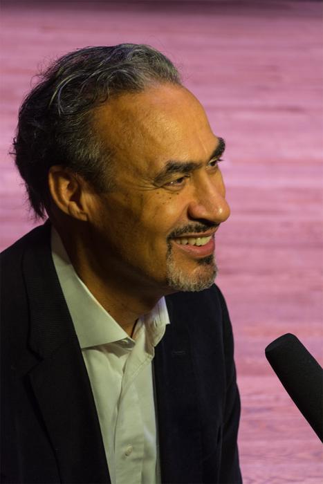 Philip Freelon at a microphone. He is wearing a suit and shirt. He is smiling and has short, styled hair and a thin goatee. A purple background sets the image. 