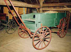 Photograph of green wooden wagon with red wooden wheels. There is what appears to be a shelf that covers a compartment in the bottom of the wagon. There is a pile of hay in the wagon, as well. 